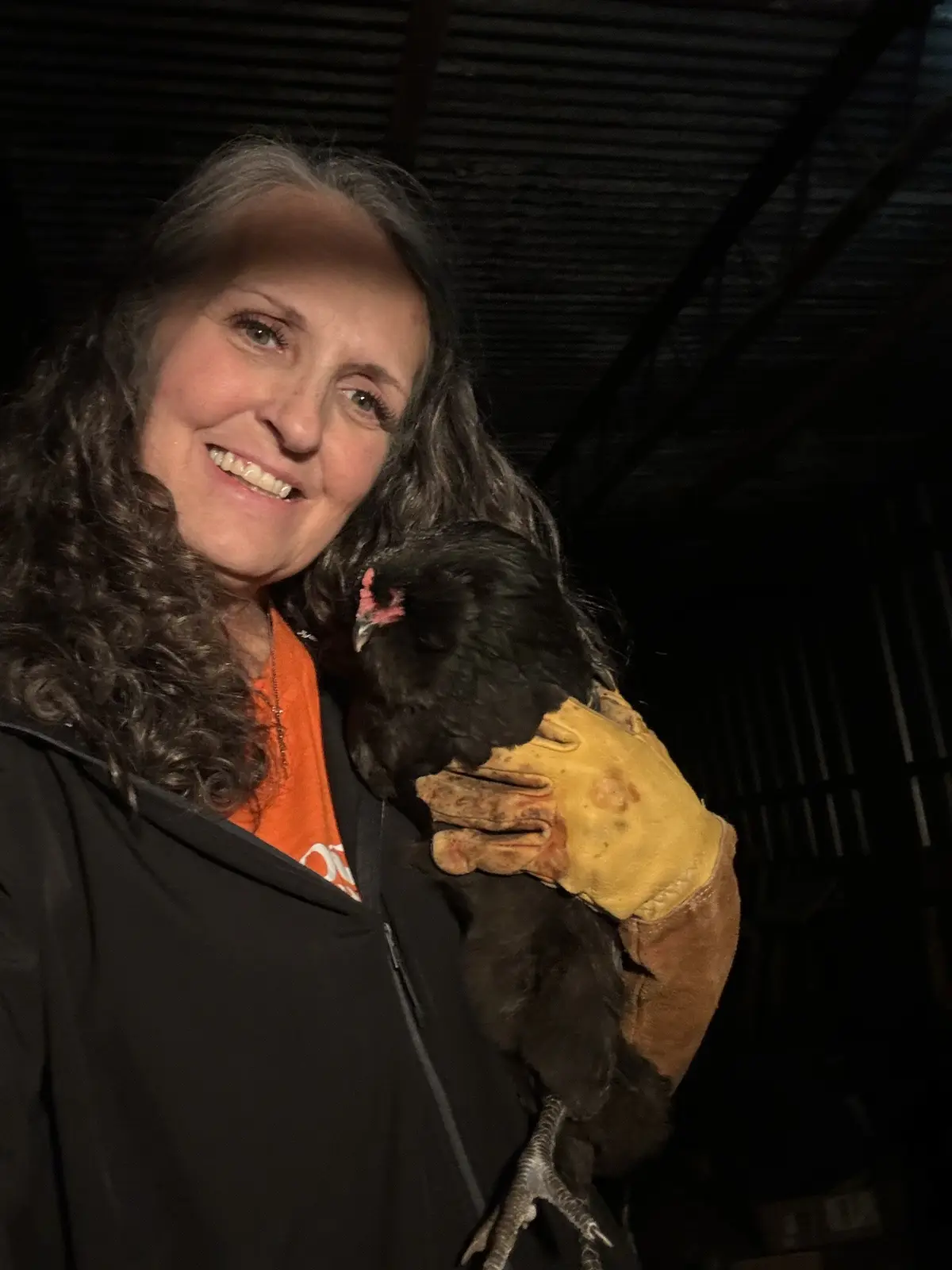 A woman with long dark hair and a warm smile holds a black chicken in her gloved hand. She wears a black jacket over an…