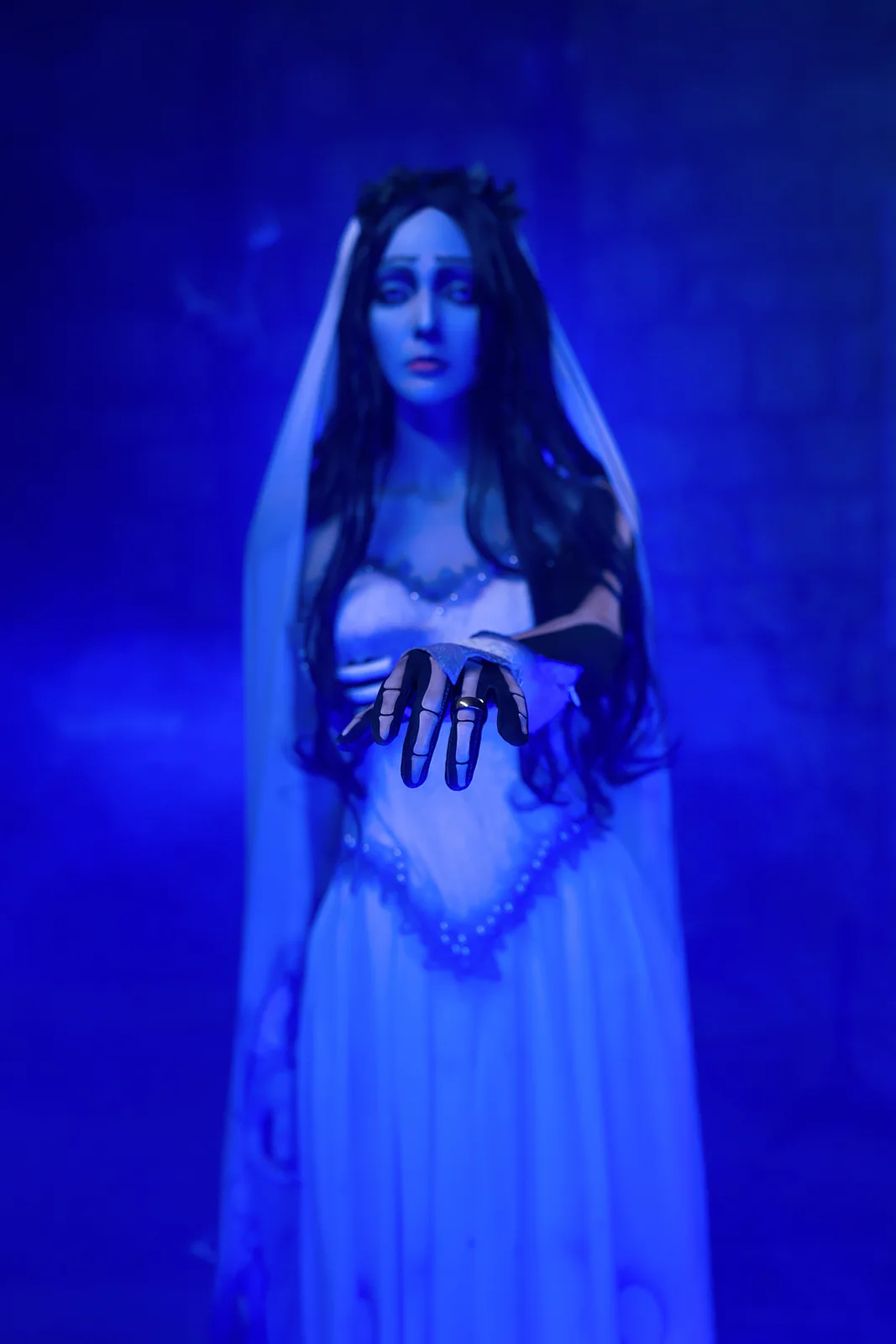 A person dressed as a ghostly bride in a white wedding gown and veil, standing in a dim blue-lit environment. Their hands…