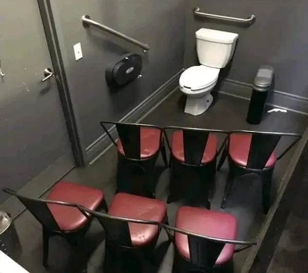 An overhead view of a small, dark gray bathroom stall containing a white toilet, grab bars, and a trash can, but also six…