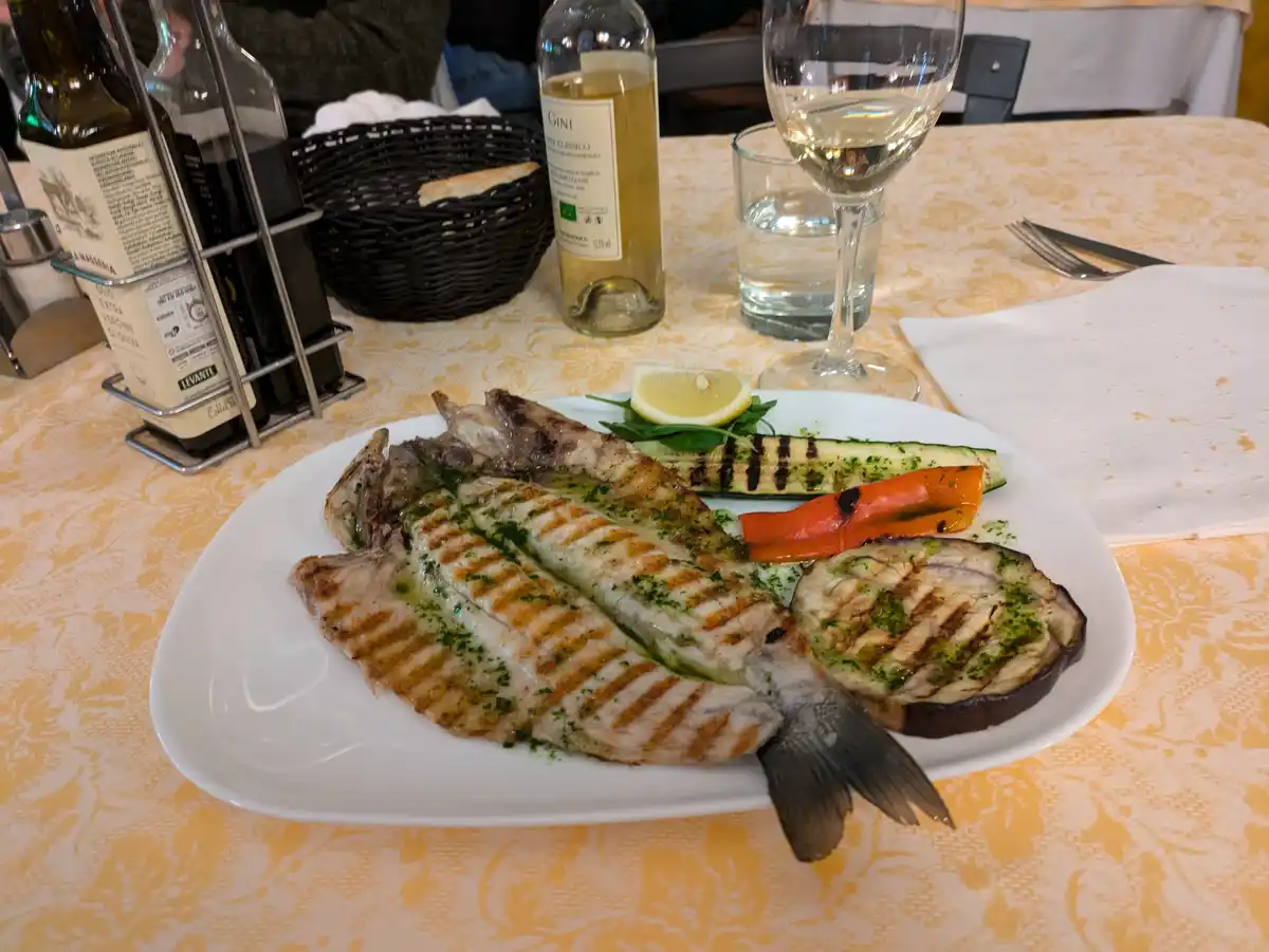 A plated grilled whole fish with visible grill marks, accompanied by grilled zucchini, eggplant, and red pepper, garnished…