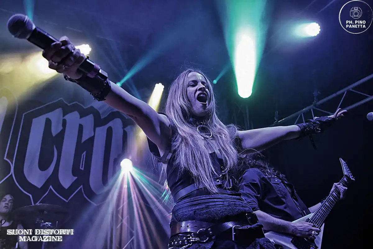 A female metal singer with long blonde hair, wearing black leather and a choker, passionately sings into a microphone with…