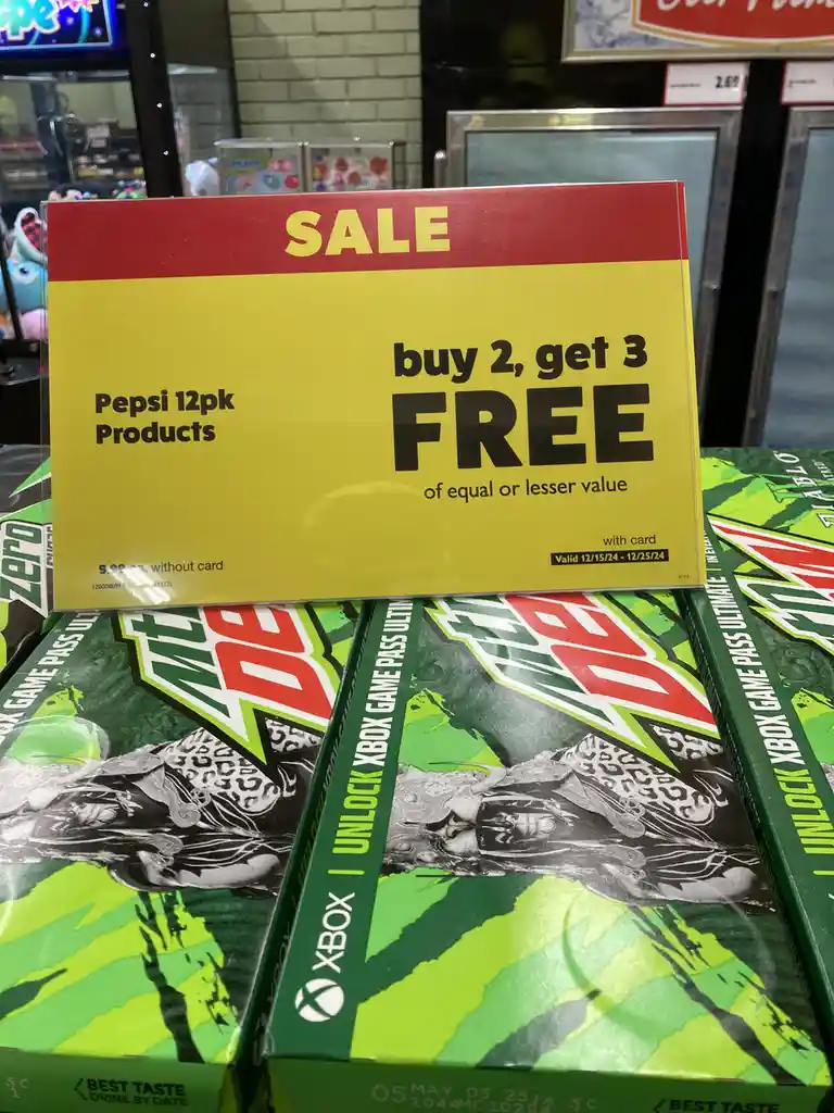 A yellow and red store sale sign advertises 'buy 2, get 3 FREE' on Pepsi 12pk products, placed atop boxes of Mountain Dew…