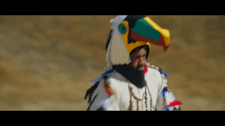 A man wearing a colorful toucan costume walks through a dry, hilly grassland. The sequence shows him starting close-up, then…