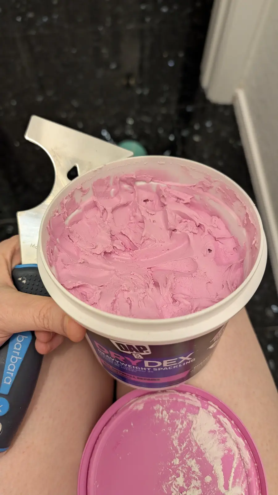 A hand holds a white tub of pink DAP Drydex heavyweight spackling compound, with a putty knife resting beside it. The pink…