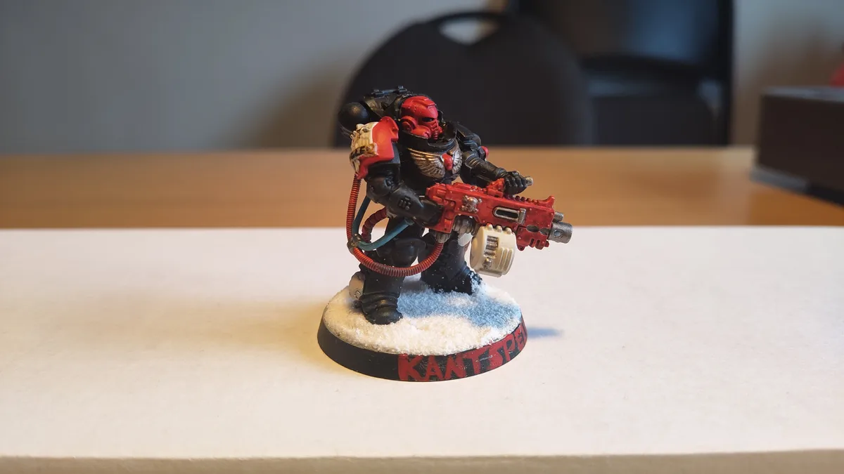 A painted miniature of a red-armored Space Marine with a red weapon stands on a snowy base labeled 'KANT SPEW'. The figure…