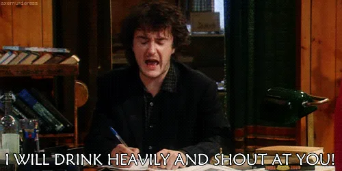The image shows a man with curly hair sitting at a wooden desk in a room with bookshelves and bottles in the background. He…