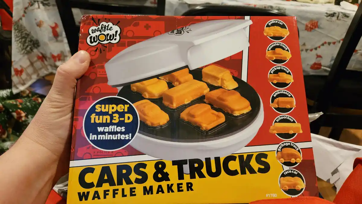 A hand holds a red and yellow box for a 'Waffle Wow! Cars & Trucks Waffle Maker'. The box shows a white appliance with six…