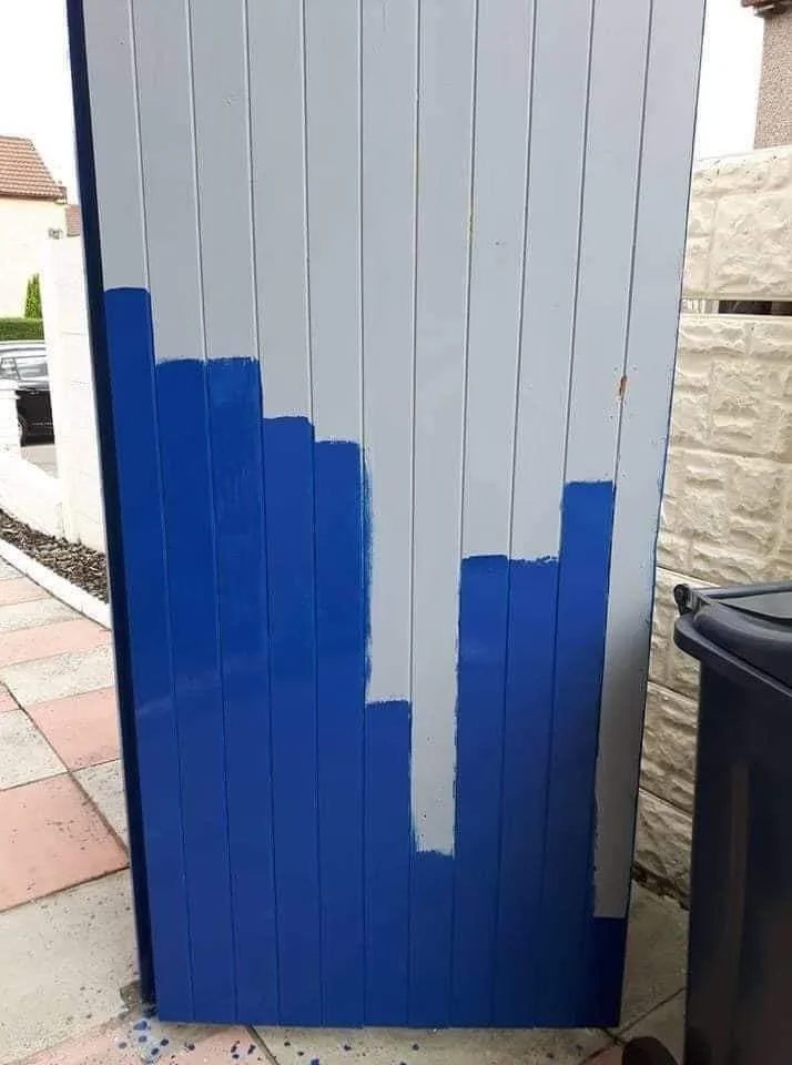 A vertical wooden fence panel is partially painted blue, with the paint applied in uneven, jagged horizontal sections…