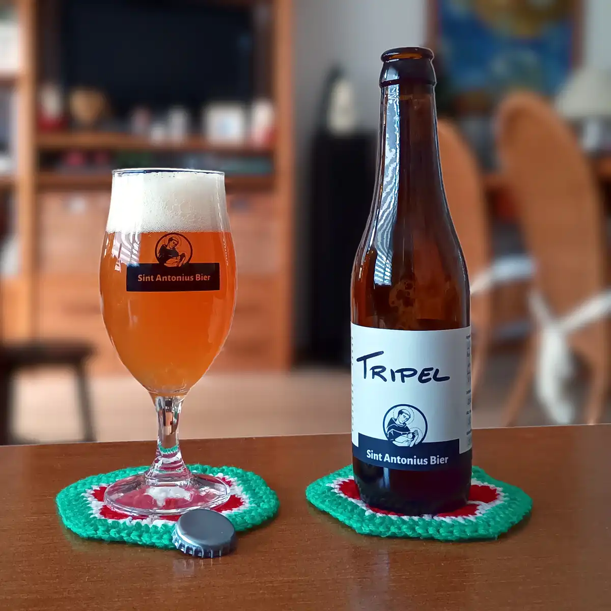 A brown beer bottle labeled 'TRIPEL' and 'Sint Antonius Bier' sits next to a glass of amber beer with the same branding…