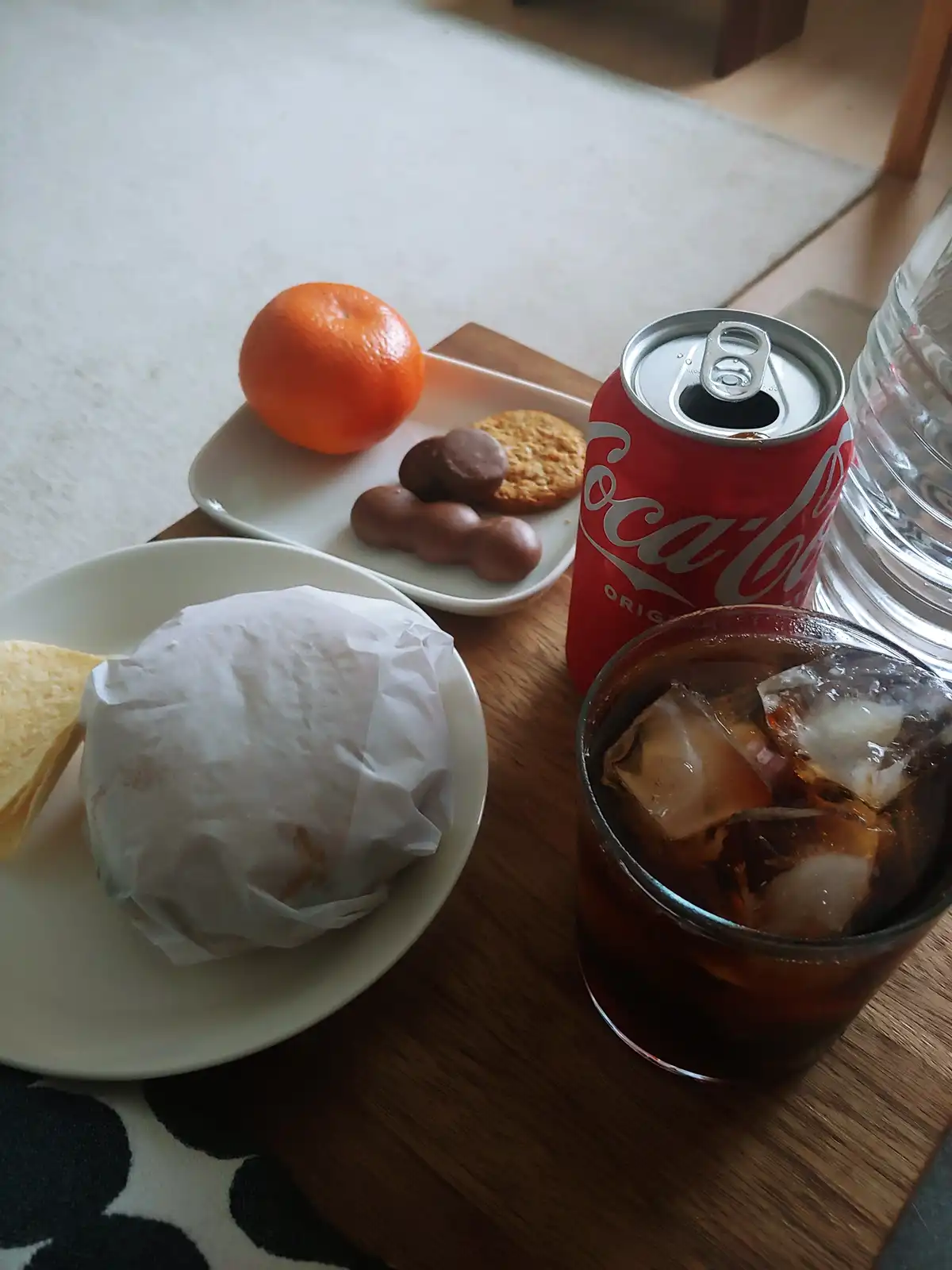 A wooden tray holds a meal including a wrapped burger, a slice of bread, a whole orange, chocolate candies, a cookie, an…