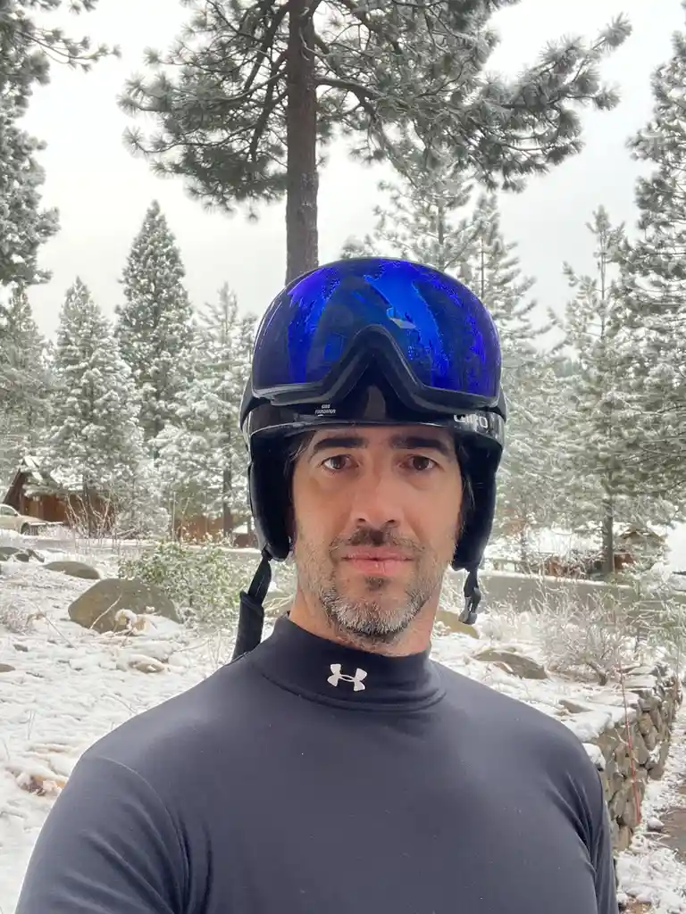 A man with a beard and mustache stands in a snowy outdoor setting with pine trees and a stone wall behind him. He wears a…
