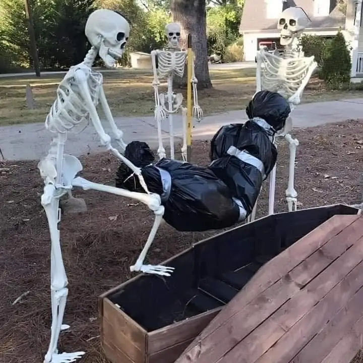 Three plastic skeleton decorations are arranged outdoors near a wooden coffin. One skeleton is seated on a black trash bag…