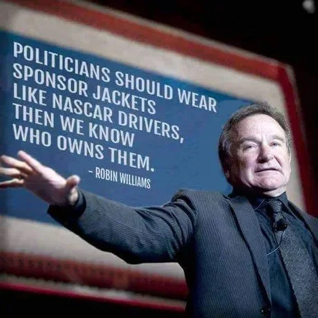 A man in a suit gestures toward a large screen displaying a quote attributed to Robin Williams that reads 'POLITICIANS…