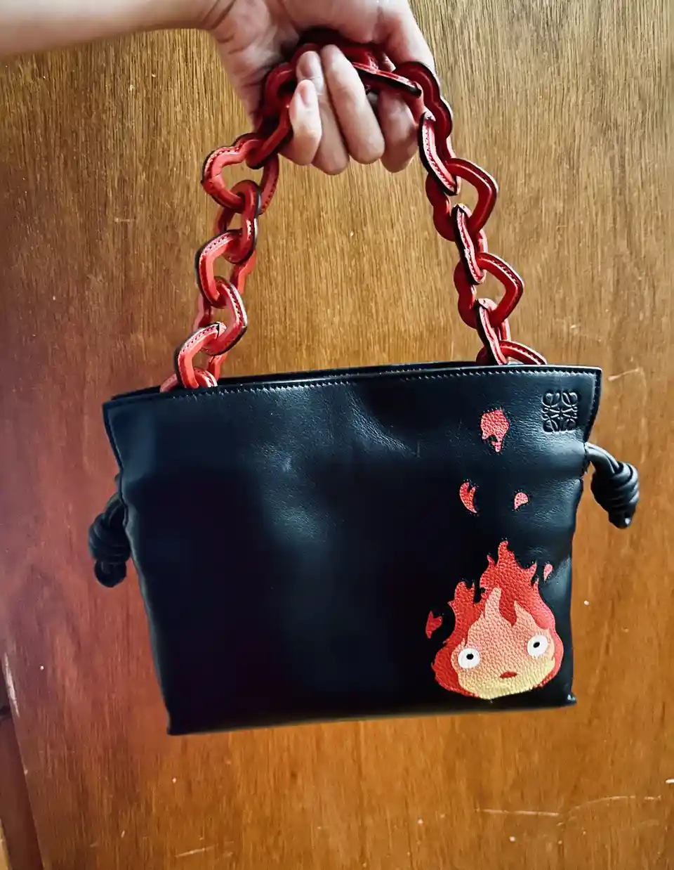A hand grips the red chain handle of a black leather purse against a wooden background. The purse features a cartoonish fire…