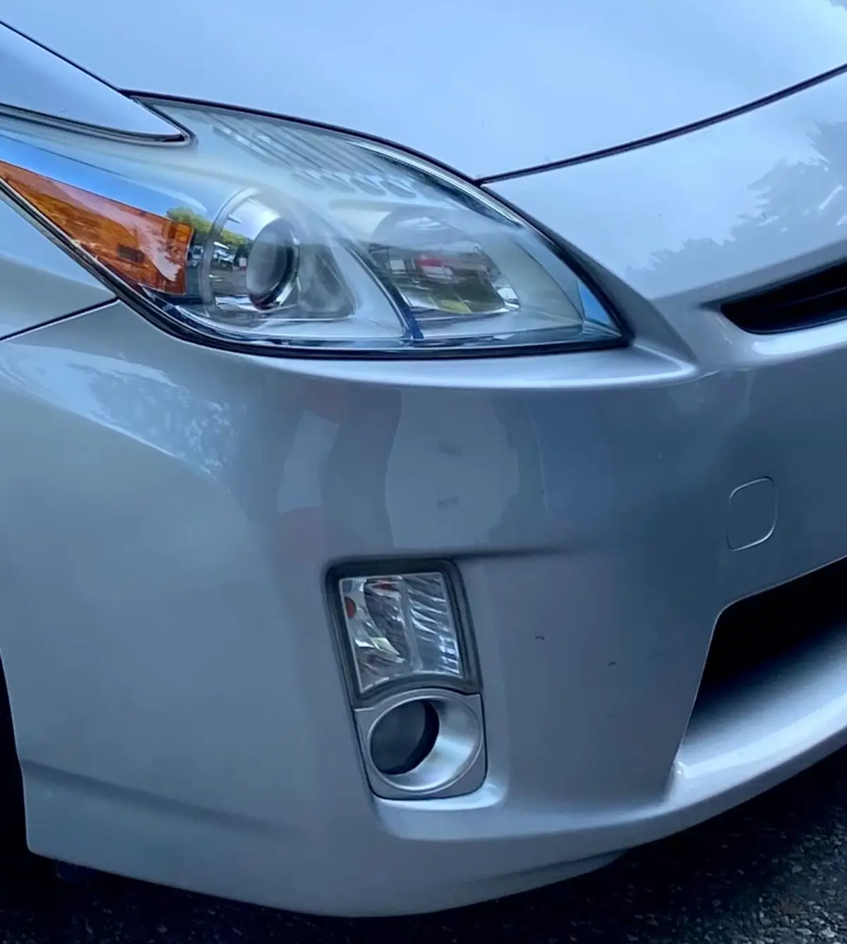 A close-up photo of the front left side of a silver car, showing its headlight, turn signal, and fog light. The car's glossy…