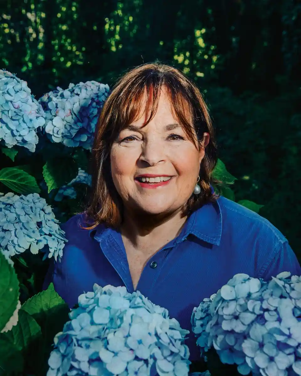 A woman with shoulder-length brown hair and a blue shirt smiles warmly while standing among large blue hydrangea blooms. The…