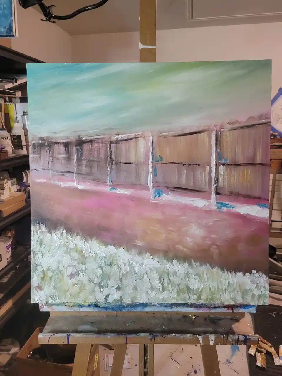 A painting on an easel in an artist's studio depicts a fence and grassy field with a surreal, dreamlike color palette of…