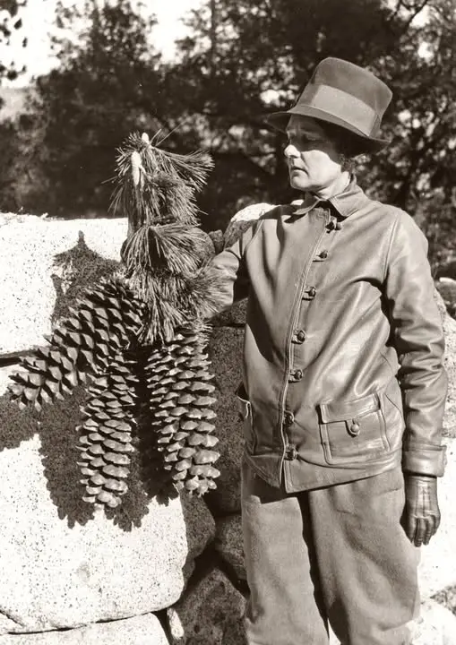 A vintage black-and-white photo shows a woman in a hat and leather jacket standing beside a large pine cone. She is holding…