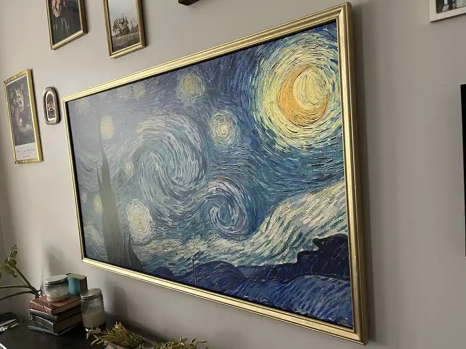 A large TV screen mounted on a wall is displaying Vincent van Gogh's 'Starry Night' painting, framed in gold. The TV is…
