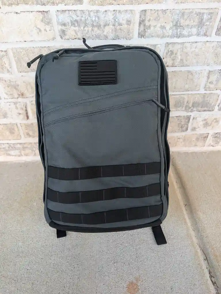 A dark gray tactical-style backpack with black MOLLE webbing and a black American flag patch sits upright against a…