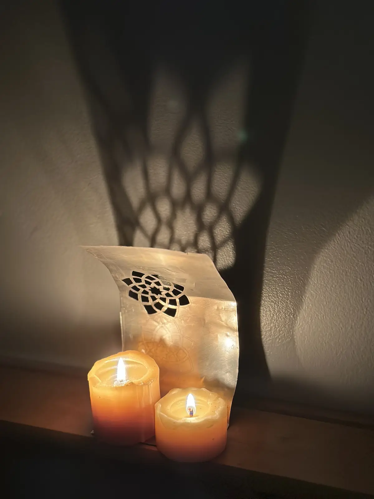 Two lit candles sit on a wooden surface next to a decorative metal candle holder with a geometric cutout pattern. The holder…