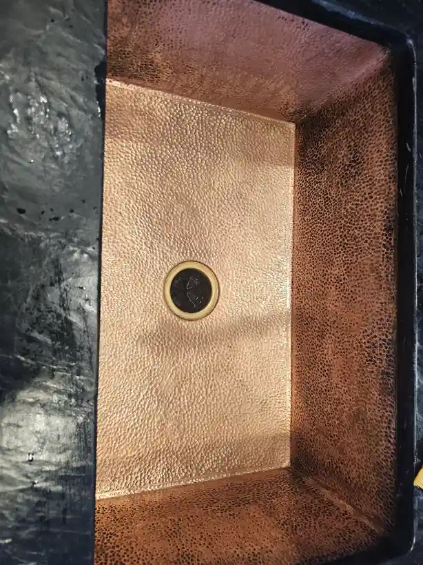 A close-up photo shows the interior of a shower stall with hammered copper walls and floor, featuring a central drain. The…