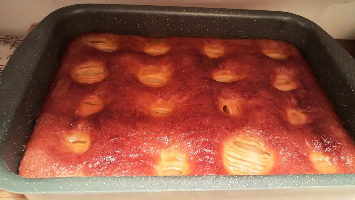 A close-up photo shows a rectangular baked good in a dark speckled baking pan, with a golden-brown, slightly cracked…
