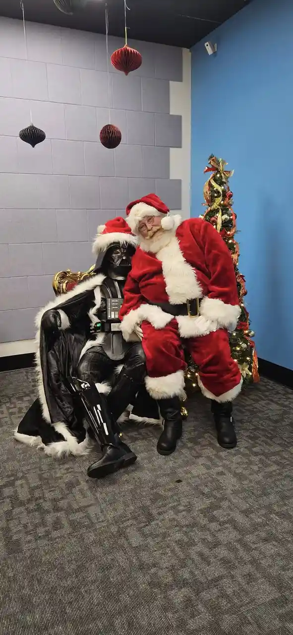 Two people dressed as Santa Claus and Darth Vader are sitting together on a throne-like chair in a room decorated for…
