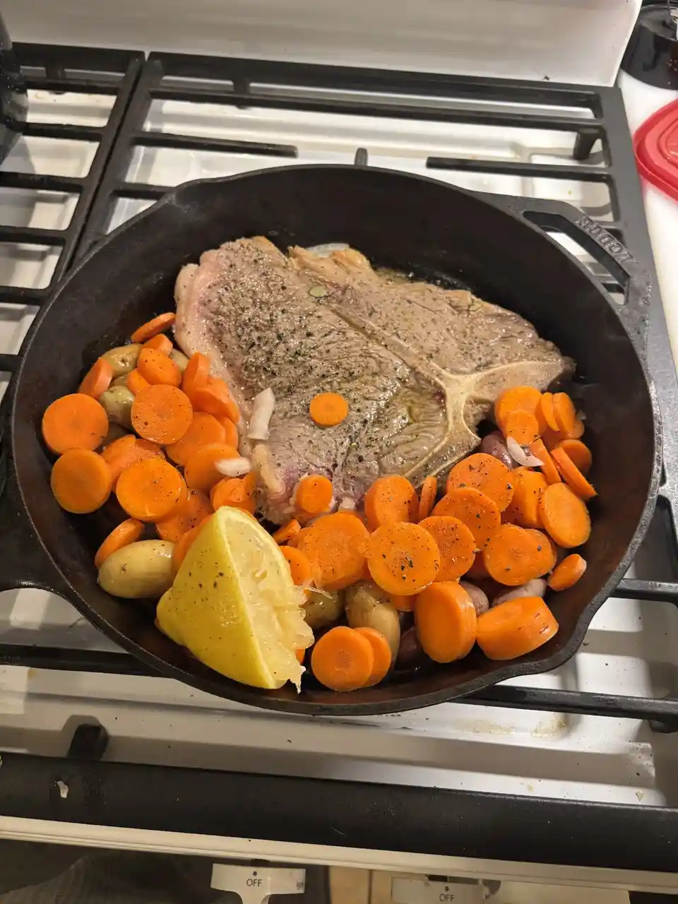 A cast iron skillet sits on a stovetop, containing a raw T-bone steak seasoned with pepper, surrounded by sliced carrots…