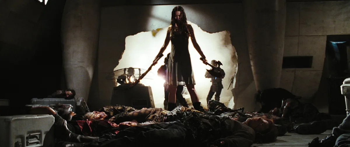 A woman in a dark dress stands holding two axes, illuminated by a bright light behind her, surrounded by bodies on the…