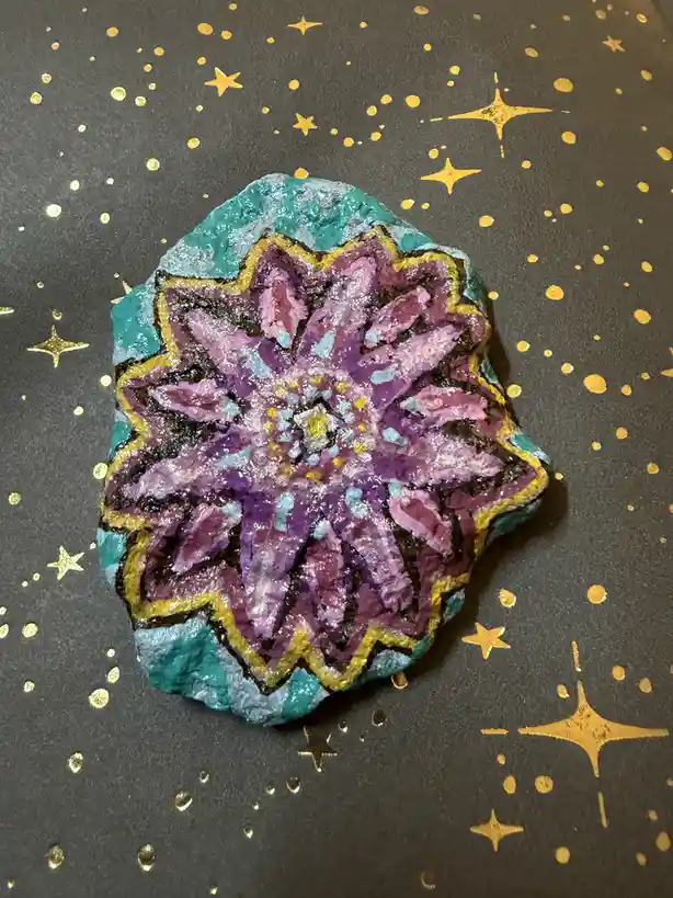 A hand-painted rock with a purple and teal mandala-like flower design sits on a dark surface decorated with gold stars and…
