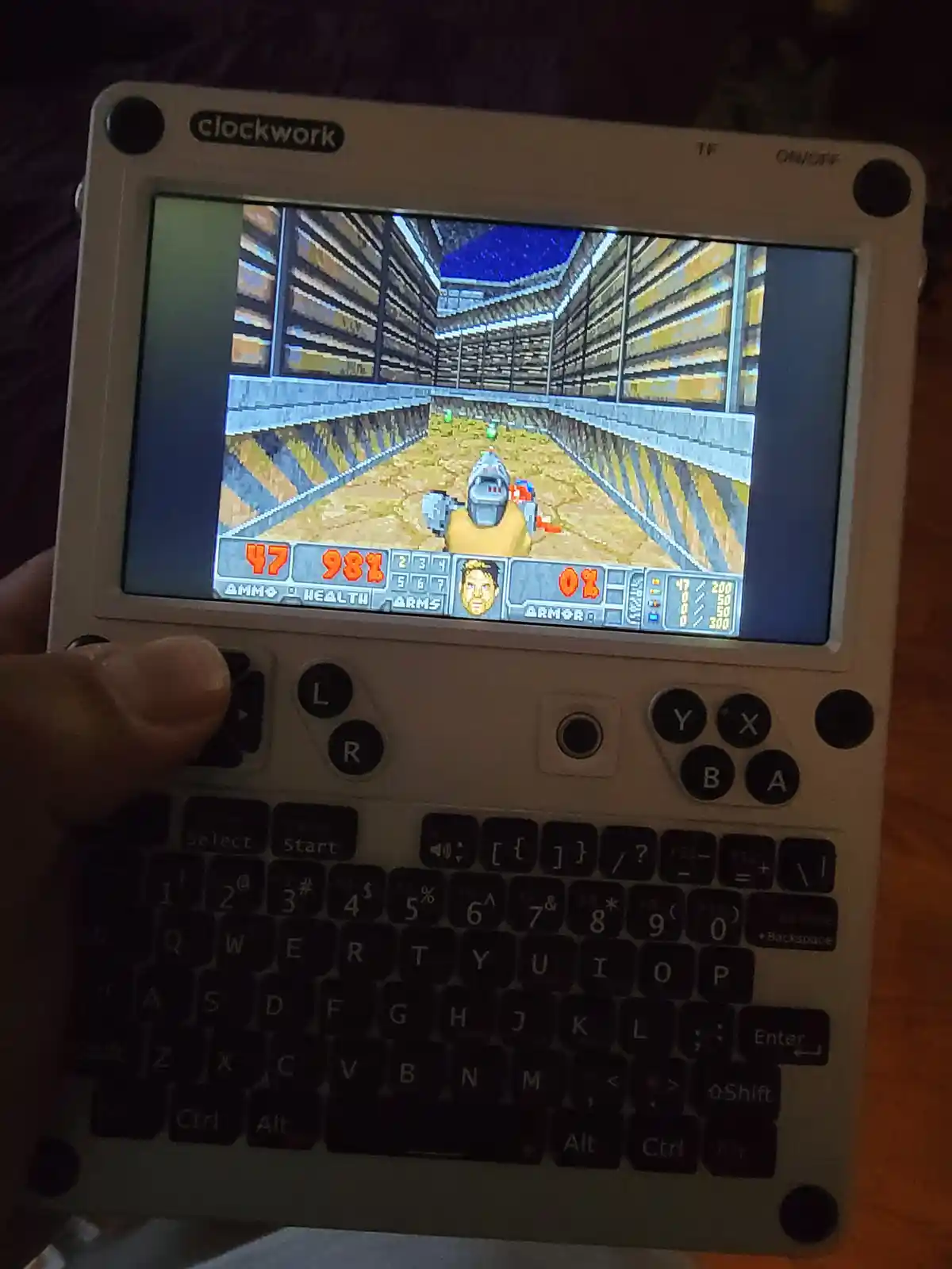 A hand holds a white 'clockwork' handheld device displaying a first-person shooter game with a health bar at 98% and armor…