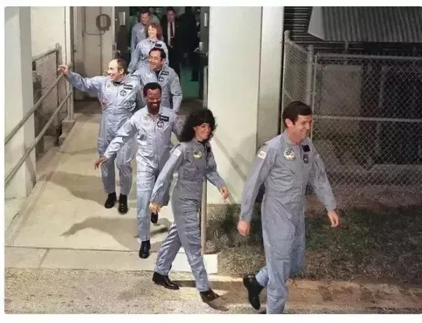 A group of astronauts in grey flight suits walk down a concrete ramp, with one woman in the center wearing a large, dark wig…