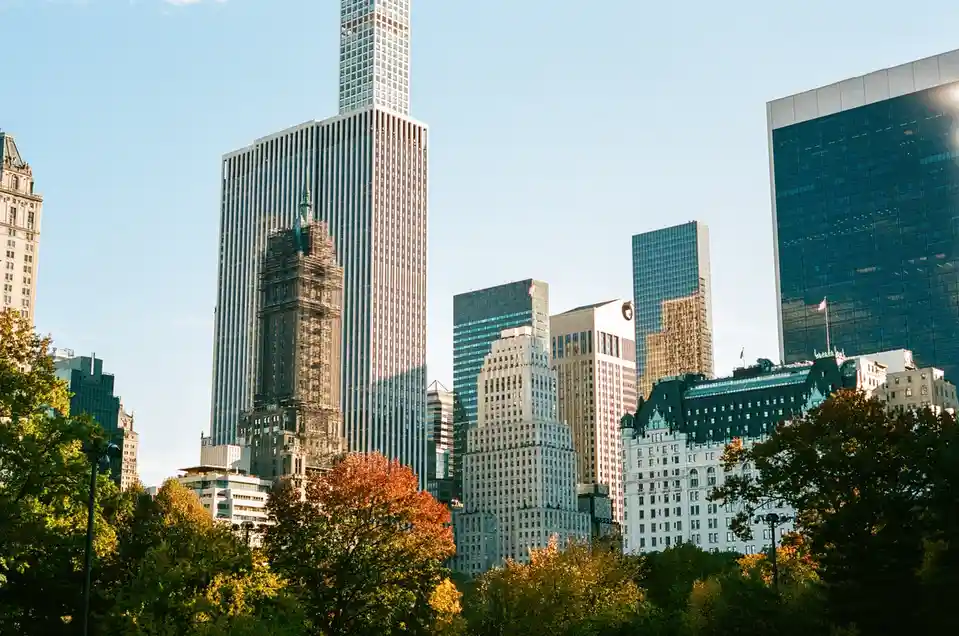 A city skyline with tall glass and steel skyscrapers under a clear blue sky, framed by trees with autumn foliage in the…
