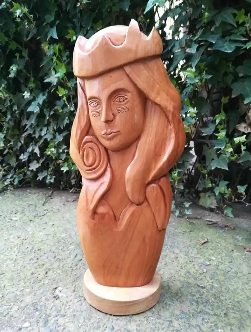 A wooden carving of a female figure with long hair, a crown, and a rose, standing on a pedestal outdoors against a backdrop…