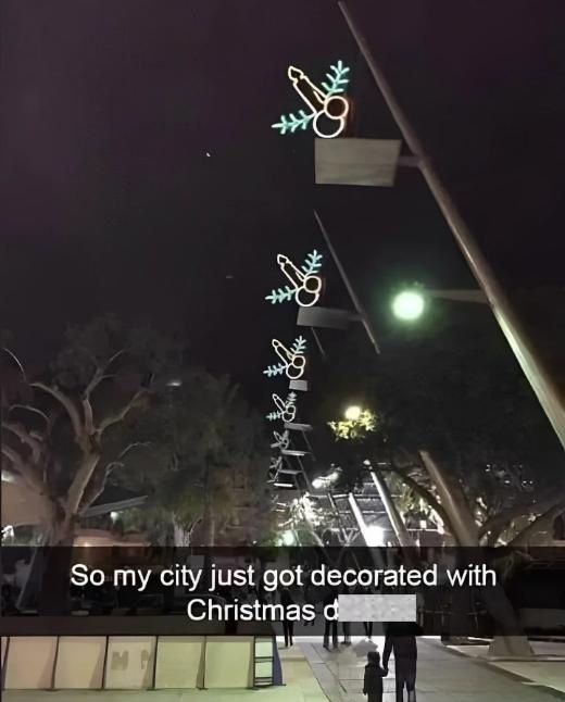 A nighttime photo shows a street decorated with unusual, glowing neon lights shaped like scissors and pine branches, hanging…