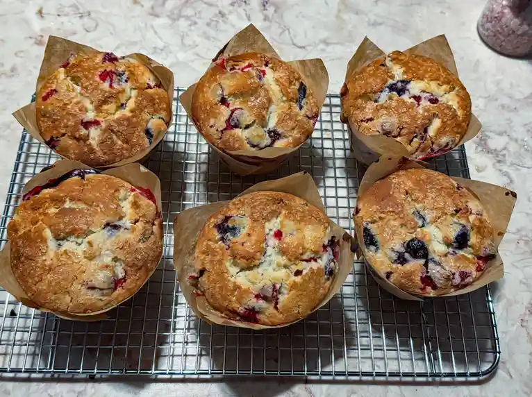 Six golden-brown berry muffins in brown paper liners sit on a metal cooling rack atop a marble countertop. The muffins have…