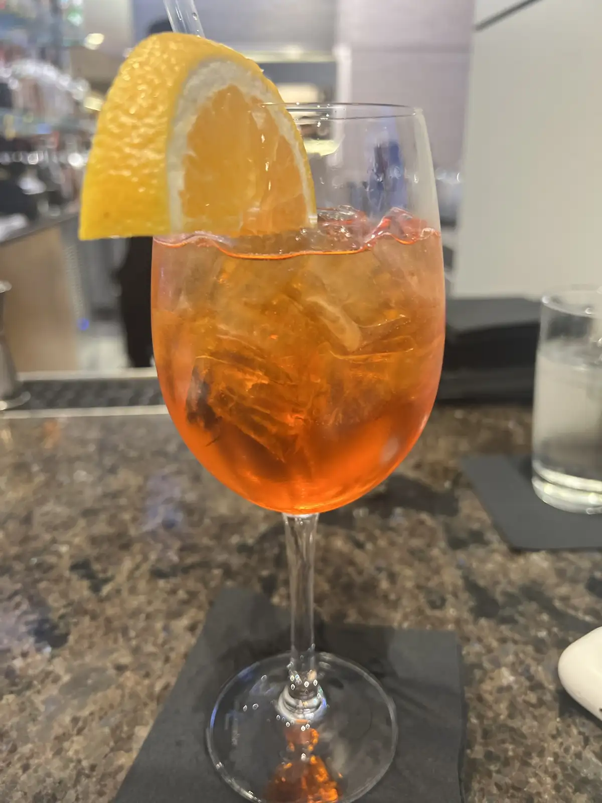 a close-up photo shows a stemmed wine glass filled with an orange-colored cocktail, ice cubes, and a lemon wedge garnish on…