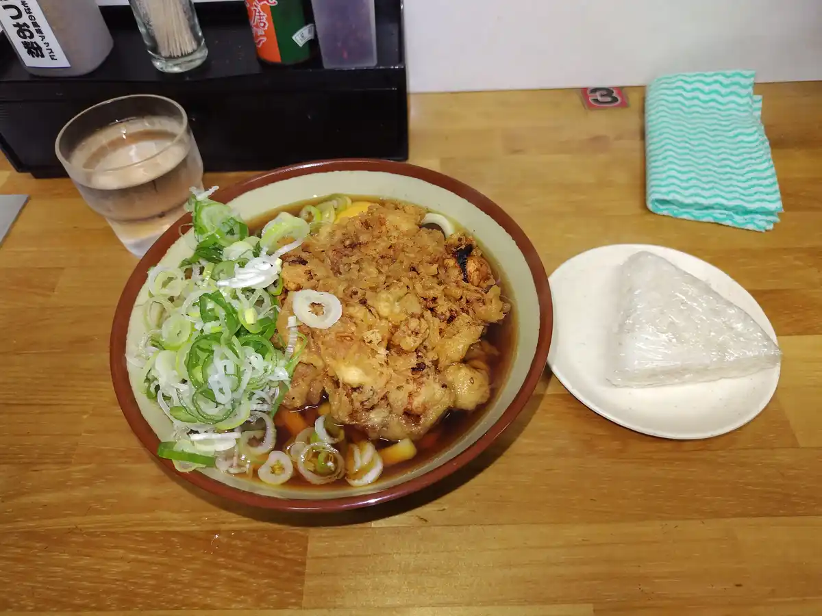 a bowl of tonkatsu ramen with crispy fried pork, sliced green onions, and broth sits on a wooden table next to a wrapped…