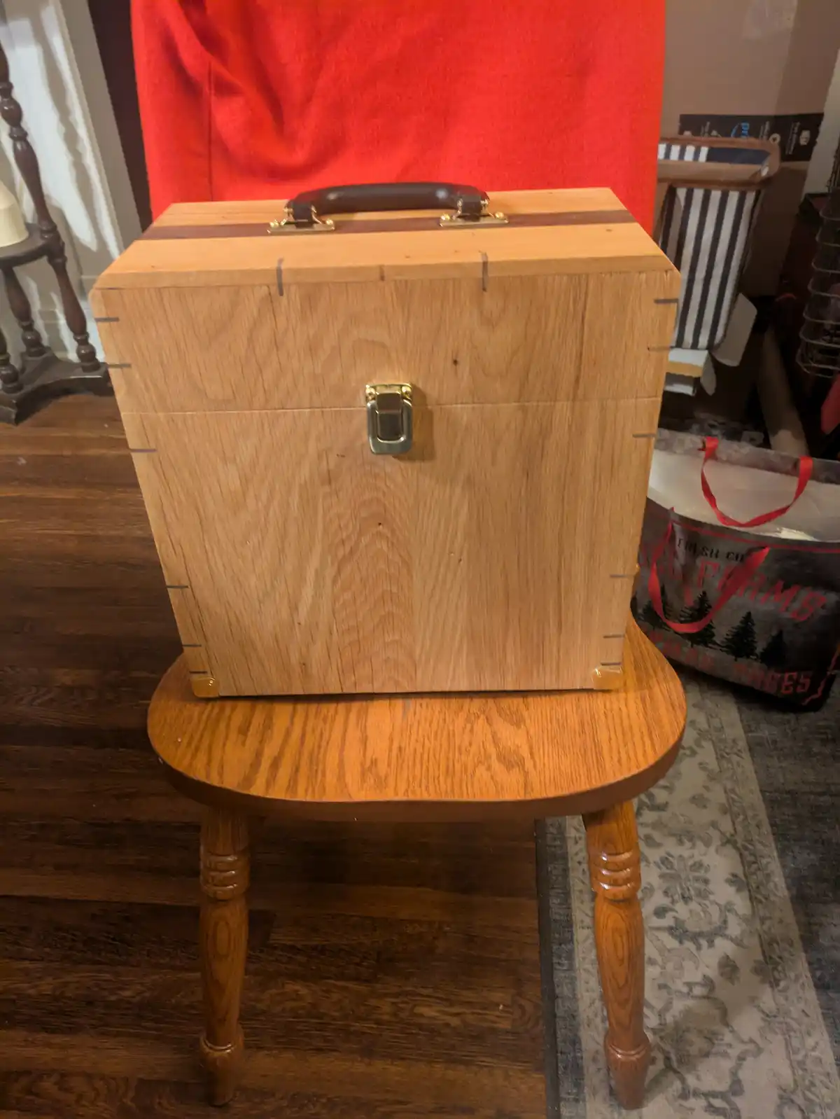 A wooden box with a handle and latch, designed to resemble a briefcase, sits atop a small wooden stool in a home setting…