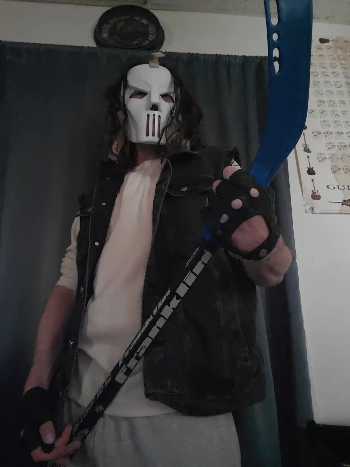 A person wearing a white skull mask with slits for eyes and mouth, long dark hair, a black denim vest over a light-colored…