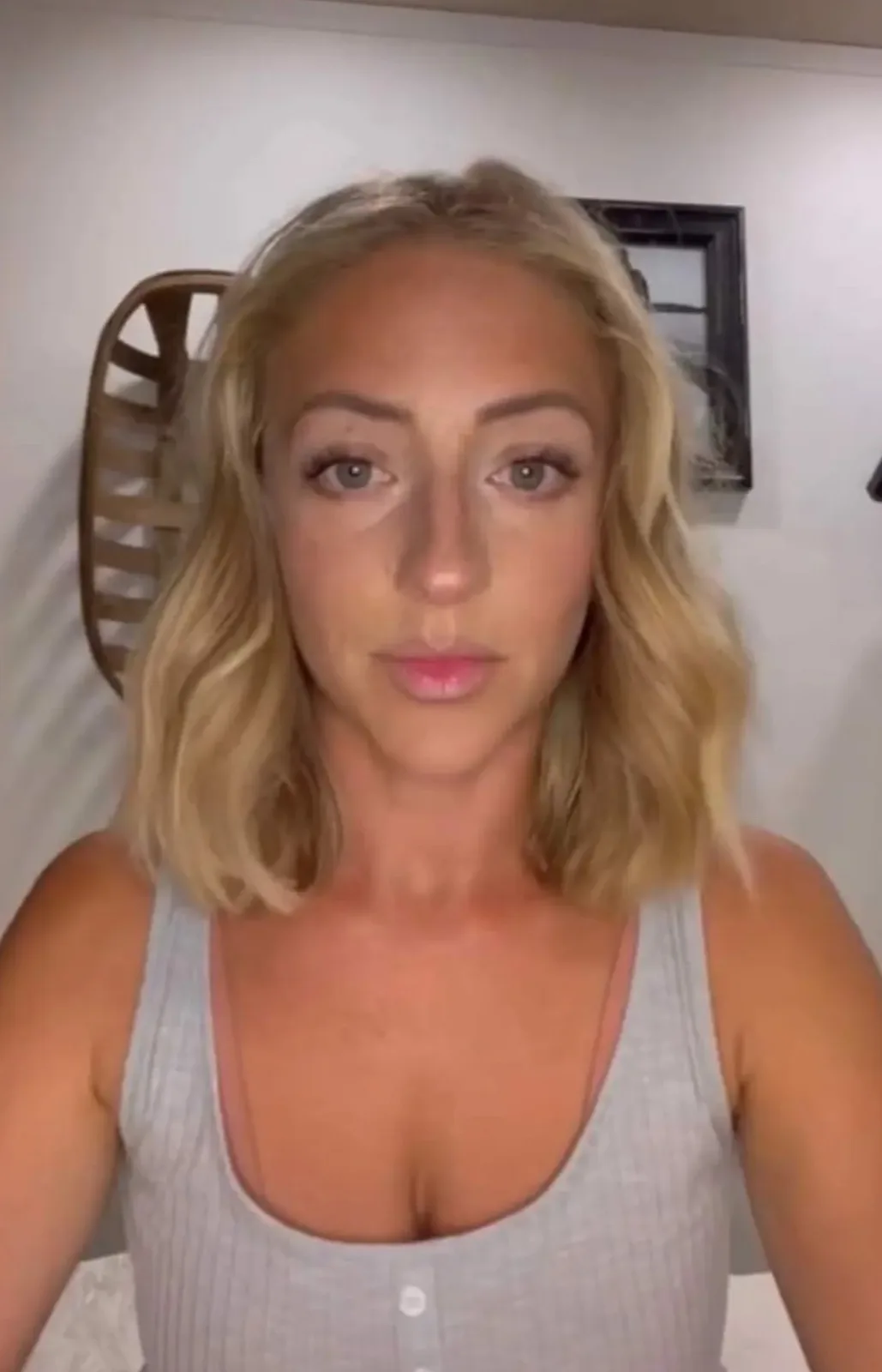 A woman with blonde wavy hair and a light gray tank top stares directly at the camera with a neutral, slightly tired…
