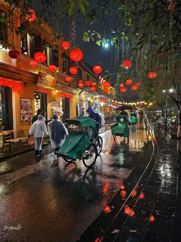 A rainy night street scene with red lanterns hanging overhead, wet pavement reflecting the lights, people in rain ponchos…