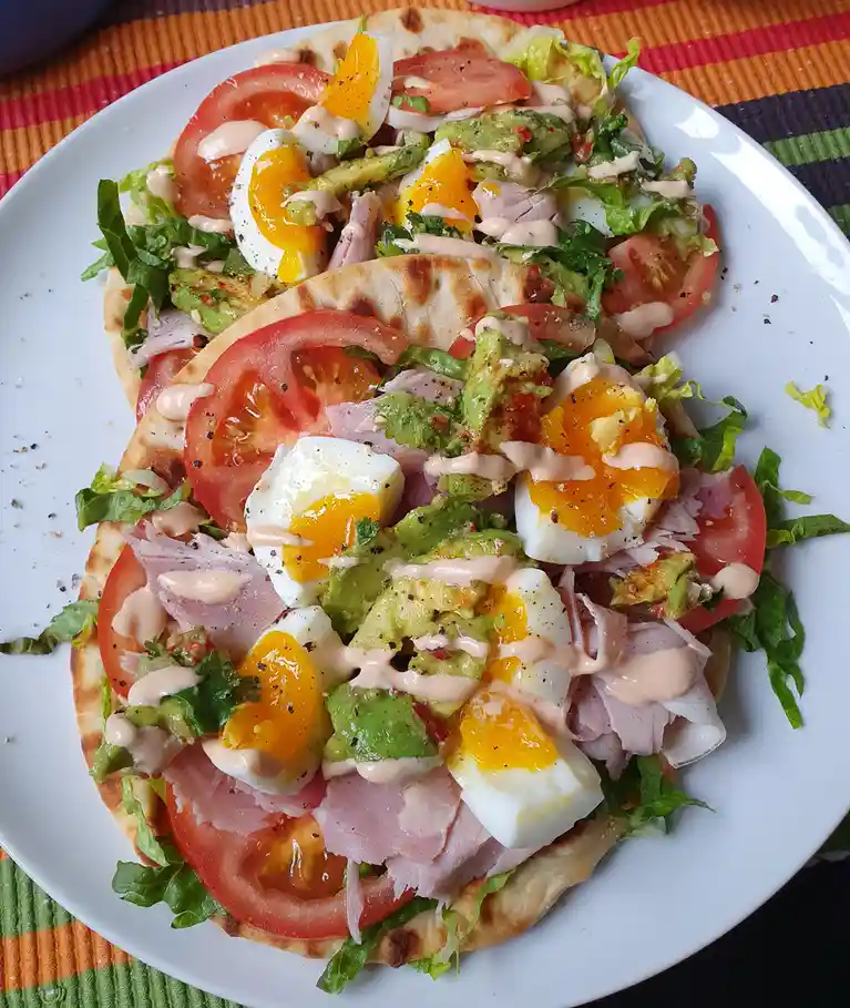 A white plate holds three overlapping flatbreads, each topped with sliced tomatoes, shredded lettuce, avocado, sliced boiled…