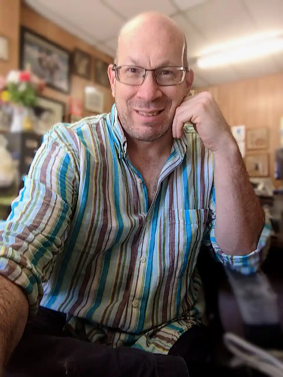 a bald man with glasses and a striped shirt smiles while resting his chin on his hand, seated in an office with framed…