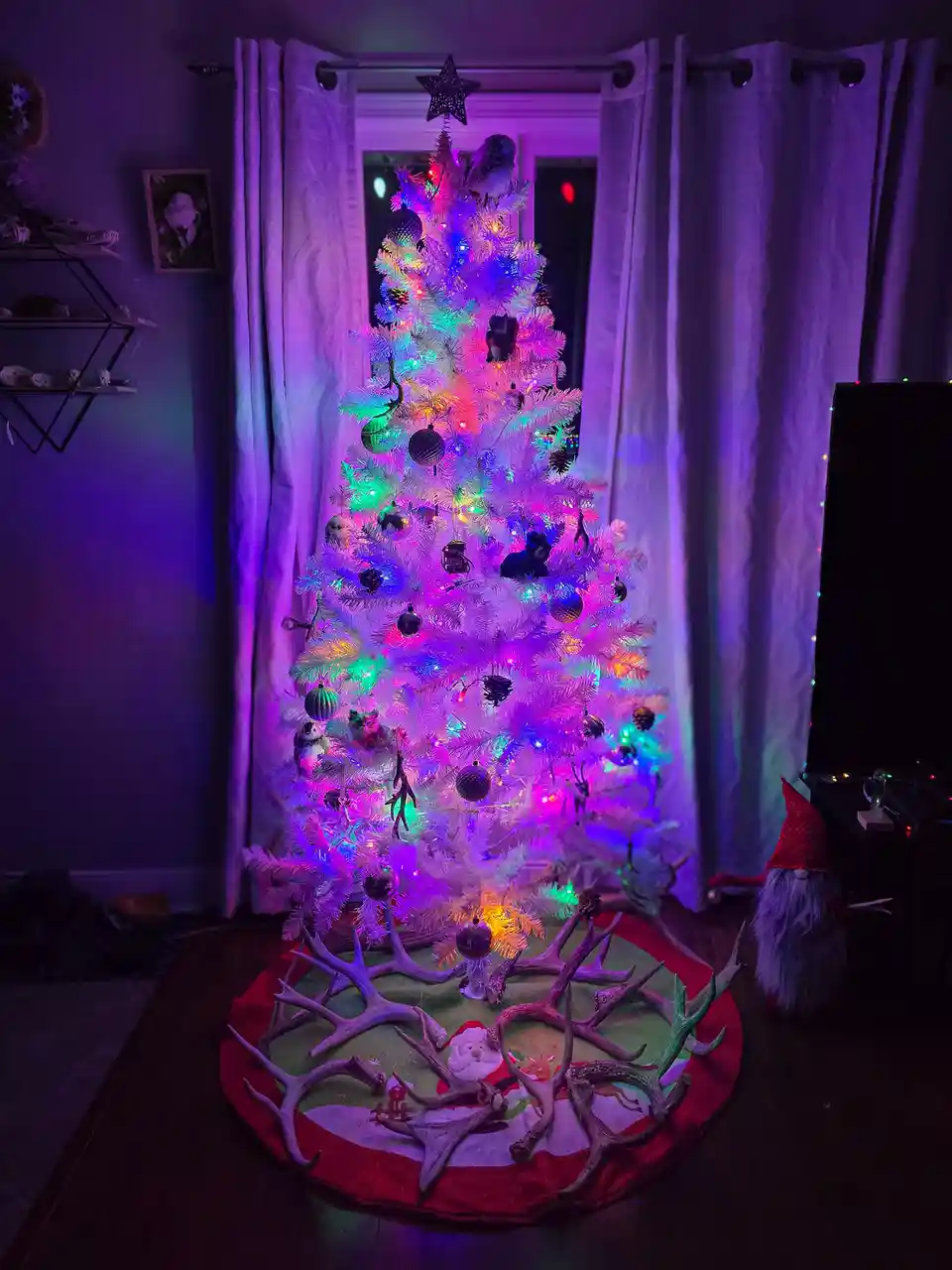 A white artificial Christmas tree is decorated with multicolored lights, ornaments, and small animal figurines, standing in…
