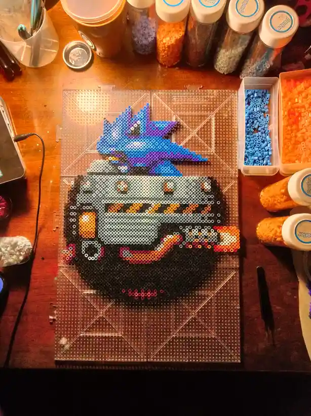 An overhead shot of a perler bead art project on a clear pegboard, depicting a stylized blue and purple dragon-like creature…