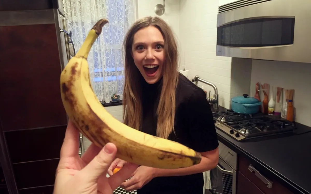 A person holds a ripe, brown-spotted banana in the foreground, while a woman in a kitchen behind them stares with wide eyes…