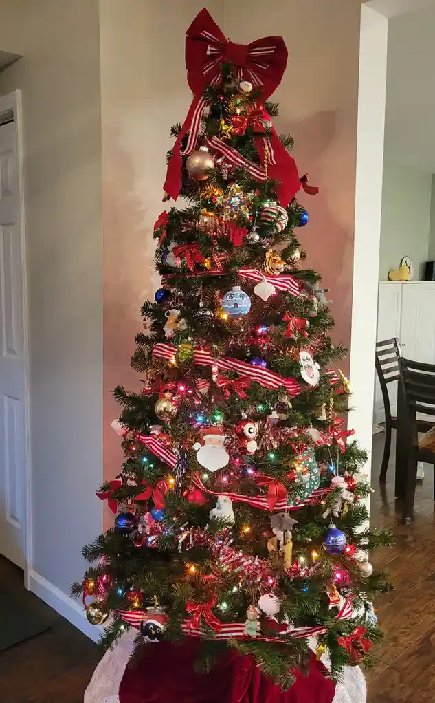 a tall, densely decorated christmas tree stands indoors, adorned with numerous red bows, colorful lights, assorted ornaments…
