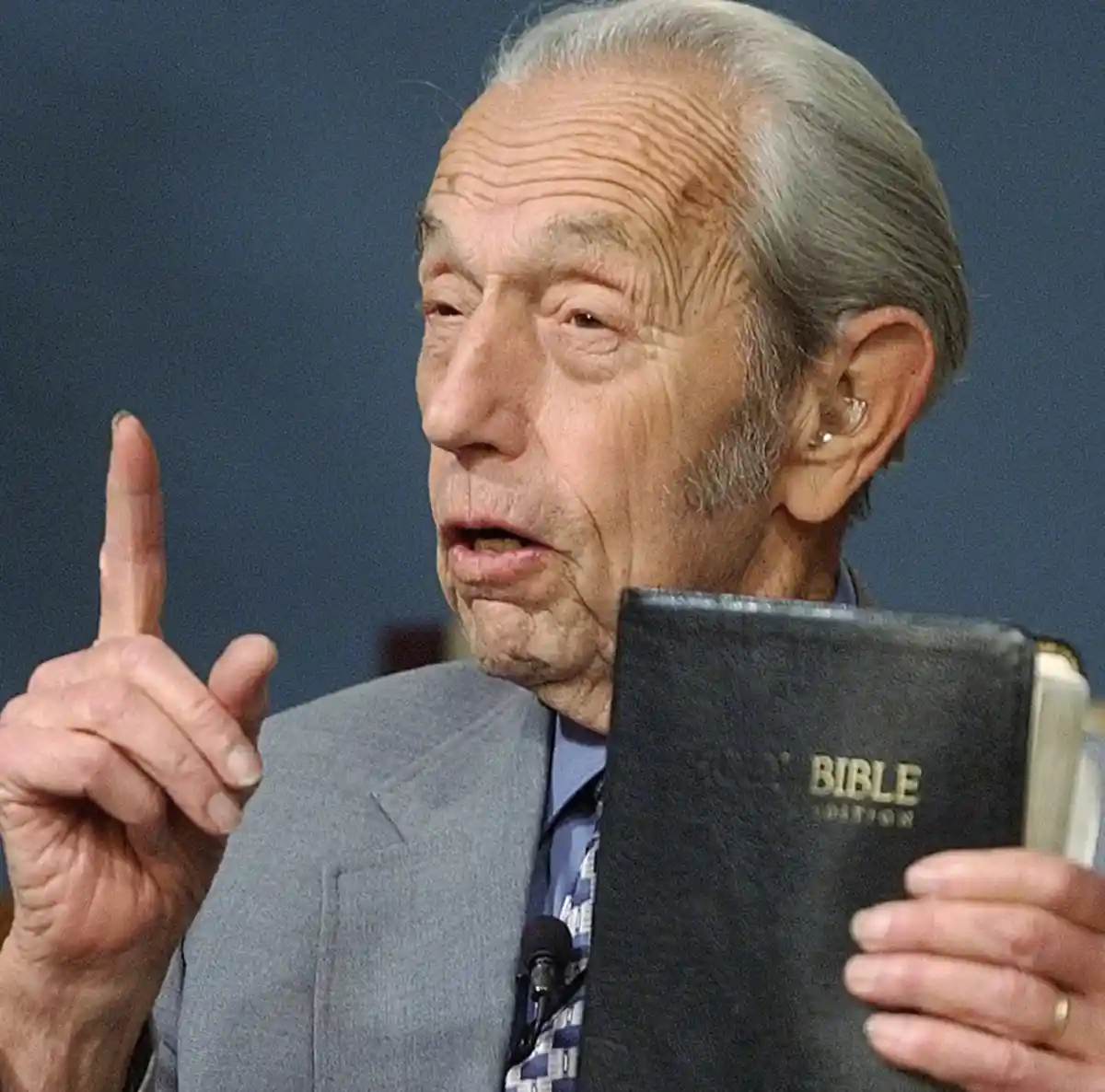 an elderly man with gray hair, wearing a gray suit and tie, holds a black Bible in his left hand while raising his right…